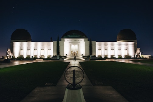 An illuminated observatory building stands against a dark night sky, its lights casting a warm glow on the white facade. The structure features a central dome flanked by two smaller domes, and a symmetrical arrangement of windows. A pathway leads up to the entrance, with neatly trimmed lawns on either side.