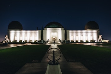 An illuminated observatory building stands against a dark night sky, its lights casting a warm glow on the white facade. The structure features a central dome flanked by two smaller domes, and a symmetrical arrangement of windows. A pathway leads up to the entrance, with neatly trimmed lawns on either side.