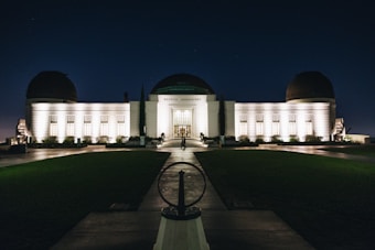 An illuminated observatory building stands against a dark night sky, its lights casting a warm glow on the white facade. The structure features a central dome flanked by two smaller domes, and a symmetrical arrangement of windows. A pathway leads up to the entrance, with neatly trimmed lawns on either side.