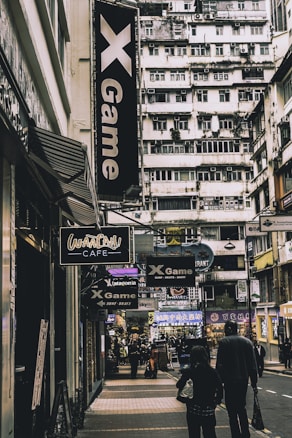 A bustling urban street scene features tall apartment buildings in the background with a variety of signs for shops and cafes in the foreground. The signs are written in different languages, including English with names like 'X Game' and 'Waaah! Cafe'. People are walking down the street, giving the scene a lively atmosphere.