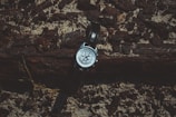 A wristwatch rests against a textured wooden and rocky backdrop. The watch has a metallic case with a dark leather strap, and it displays a detailed face with various small dials.