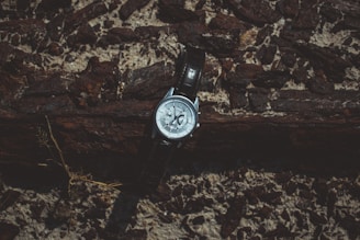A wristwatch rests against a textured wooden and rocky backdrop. The watch has a metallic case with a dark leather strap, and it displays a detailed face with various small dials.