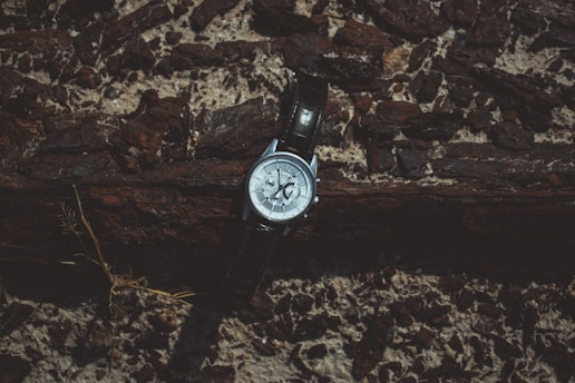 A wristwatch rests against a textured wooden and rocky backdrop. The watch has a metallic case with a dark leather strap, and it displays a detailed face with various small dials.