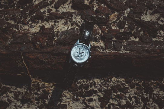 A wristwatch rests against a textured wooden and rocky backdrop. The watch has a metallic case with a dark leather strap, and it displays a detailed face with various small dials.