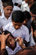 A group of children in uniform gather closely as one child receives a face painting of a national flag on his cheek by an adult using a small paintbrush.
