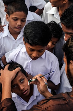 A group of children in uniform gather closely as one child receives a face painting of a national flag on his cheek by an adult using a small paintbrush.