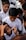 A group of children in uniform gather closely as one child receives a face painting of a national flag on his cheek by an adult using a small paintbrush.