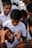 A group of children in uniform gather closely as one child receives a face painting of a national flag on his cheek by an adult using a small paintbrush.