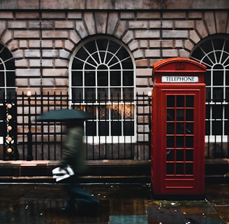 time lapse photography of woman walking on street while holding umbrella near London telephone booth beside wall