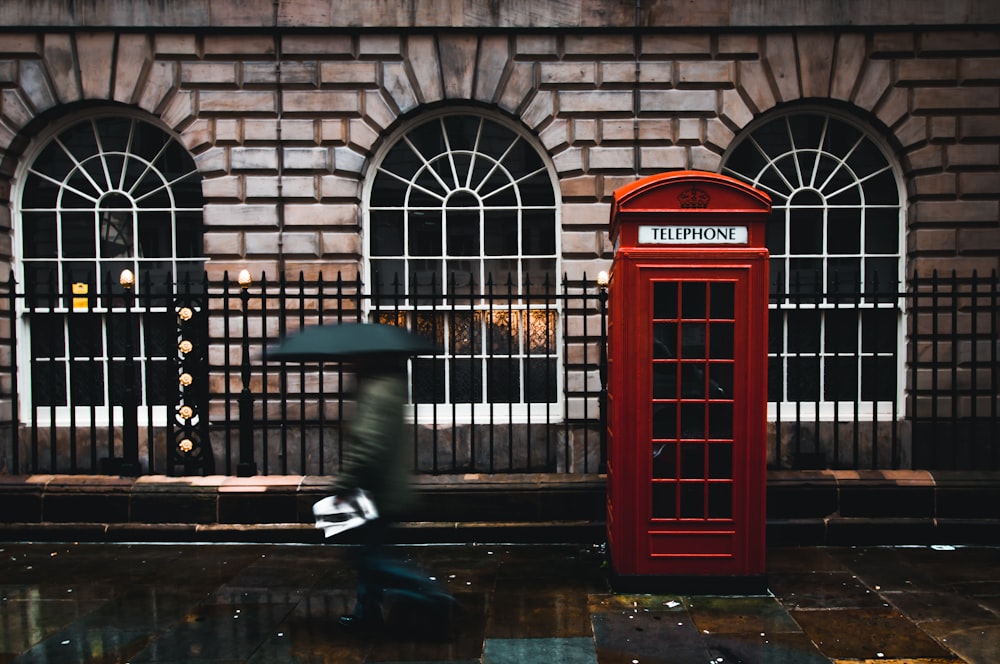 time lapse photography of woman walking on street while holding umbrella near London telephone booth beside wall