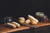 A variety of pastries are arranged on a wooden board, including éclairs and cream puffs with various toppings. There is a jar of blueberries and two small jars, one possibly containing cocoa. A fork and knife are placed next to the pastries, and the background is dark, adding a cozy, inviting atmosphere.