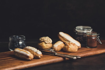A variety of pastries are arranged on a wooden board, including éclairs and cream puffs with various toppings. There is a jar of blueberries and two small jars, one possibly containing cocoa. A fork and knife are placed next to the pastries, and the background is dark, adding a cozy, inviting atmosphere.