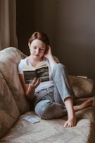 A person is sitting comfortably on a textured sofa, engrossed in reading a book titled 'THE SHACK' by Wm. Paul Young. Dressed in a white t-shirt and jeans, their posture is relaxed with one knee bent and the other leg stretched out. An object that appears to be a phone is placed beside them on the couch.