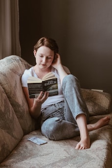 A person is sitting comfortably on a textured sofa, engrossed in reading a book titled 'THE SHACK' by Wm. Paul Young. Dressed in a white t-shirt and jeans, their posture is relaxed with one knee bent and the other leg stretched out. An object that appears to be a phone is placed beside them on the couch.