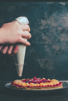 A person is decorating a fruit tart with a piping bag filled with cream or icing. The tart is garnished with a variety of fruits including raspberries, orange slices, and red currants. The background is a dark, textured surface that contrasts with the colorful fruit.