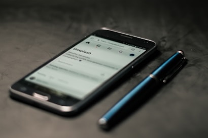 A smartphone displaying a web page is placed on a dark surface next to a blue pen. The phone screen shows a website with text and icons visible.