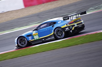 A blue race car with the number 99 on the side is speeding on a track, showing motion blur. The vehicle is adorned with various sponsor logos including PRO and DHL. It features a prominent rear wing and sleek aerodynamic design. The race track surface is visible along with red and white lines on the side.