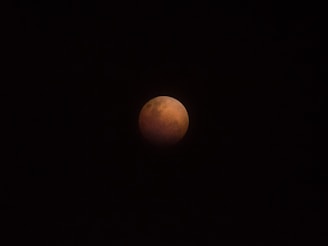 A celestial event captures the Earth's shadow covering the moon, giving it a reddish hue known as a blood moon, set against a black night sky.
