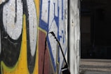 An electric guitar rests against a wall adorned with vibrant street art. The graffiti features bold colors like yellow, blue, and black, creating an urban and artistic atmosphere. The scene is set outdoors, possibly in an urban alley or a street corner, with cobblestone pavement visible and a dark, moody background.