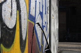An electric guitar rests against a wall adorned with vibrant street art. The graffiti features bold colors like yellow, blue, and black, creating an urban and artistic atmosphere. The scene is set outdoors, possibly in an urban alley or a street corner, with cobblestone pavement visible and a dark, moody background.
