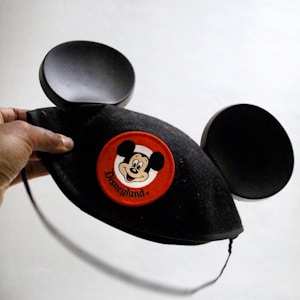 A hand is holding a classic Disneyland Mickey Mouse ear hat. The hat is black with large round ears and features a red patch with the smiling face of a famous cartoon character and the word 'Disneyland' beneath it.