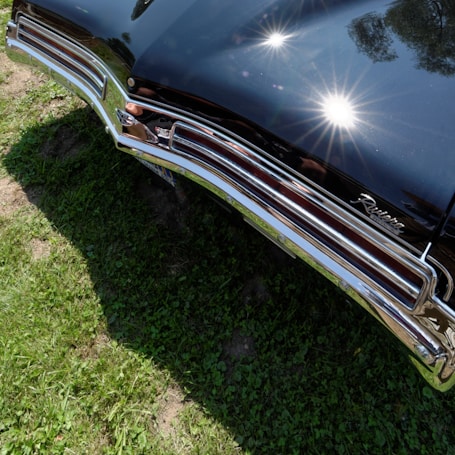 A close-up of a classic car’s rear end with shiny chrome detailing and a nameplate that reads 'Riviera.' The car's black exterior reflects the sunlight and a tree silhouette. The car is parked on a grassy area with some patches of exposed soil.