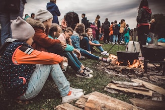 A group of children are sitting on the ground near a campfire, dressed in casual outdoor clothing. Some are wearing beanies and jackets, indicating cool weather. They appear engaged and relaxed, with logs and cooking pots over the fire. In the background, additional people are gathered, creating a lively atmosphere.
