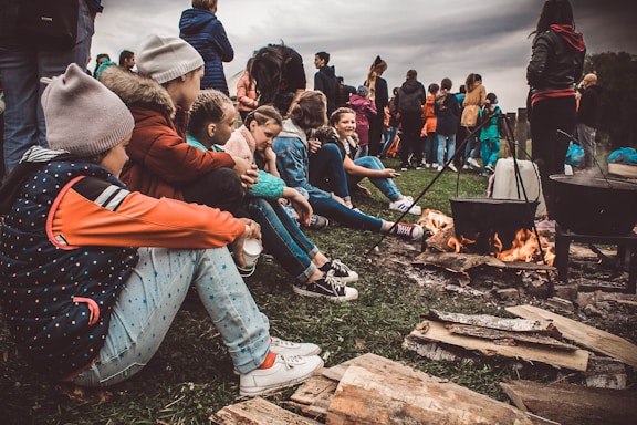 A group of children are sitting on the ground near a campfire, dressed in casual outdoor clothing. Some are wearing beanies and jackets, indicating cool weather. They appear engaged and relaxed, with logs and cooking pots over the fire. In the background, additional people are gathered, creating a lively atmosphere.