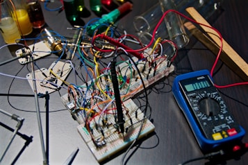 A collection of electronic components and test equipment spread across a table. There are breadboards with numerous colorful wires connected, a multimeter, and various test tubes in the background. The setup suggests a workbench for electronics or a science experiment.