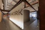 A modern co-working space with industrial design elements, featuring a cylindrical meeting room surrounded by frosted glass walls. The area is lit by soft overhead lights and has exposed ducts and beams on the ceiling. A hallway extends from the central space, showing a buzz of activity with blurred figures indicating movement.