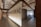 A modern co-working space with industrial design elements, featuring a cylindrical meeting room surrounded by frosted glass walls. The area is lit by soft overhead lights and has exposed ducts and beams on the ceiling. A hallway extends from the central space, showing a buzz of activity with blurred figures indicating movement.