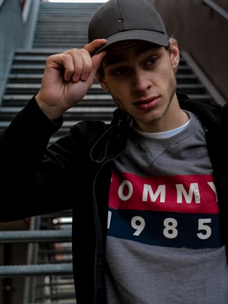 A young individual wearing a grey cap and a black jacket over a sweatshirt with the text 'TOMMY 1985'. The background consists of a staircase.