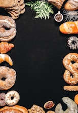 An assortment of baked goods, including bread, croissants, donuts, and pretzels, is arranged around the borders of a dark background. There are decorative elements such as green leaves and a small cup of coffee beans. The breads have different shapes, textures, and some are topped with seeds.