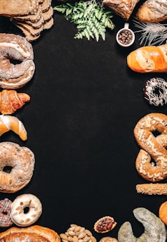 An assortment of baked goods, including bread, croissants, donuts, and pretzels, is arranged around the borders of a dark background. There are decorative elements such as green leaves and a small cup of coffee beans. The breads have different shapes, textures, and some are topped with seeds.