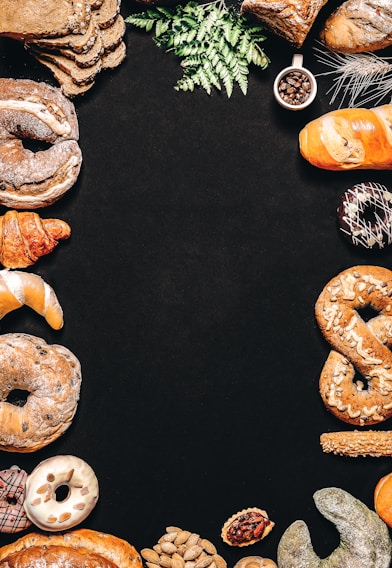 An assortment of baked goods, including bread, croissants, donuts, and pretzels, is arranged around the borders of a dark background. There are decorative elements such as green leaves and a small cup of coffee beans. The breads have different shapes, textures, and some are topped with seeds.