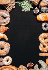 An assortment of baked goods, including bread, croissants, donuts, and pretzels, is arranged around the borders of a dark background. There are decorative elements such as green leaves and a small cup of coffee beans. The breads have different shapes, textures, and some are topped with seeds.