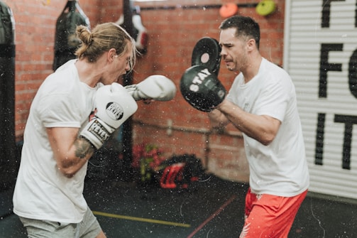 Two individuals are engaged in a boxing training session inside a gym. One person is wearing white boxing gloves and is punching toward the other, who is holding up black focus mitts. Both are dressed casually in sports attire, surrounded by punching bags and gym equipment with a brick wall background.