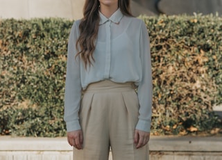 woman wearing gray button-up long-sleeved shirt and brown pants standing in front of bushes