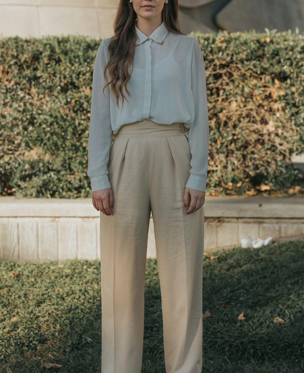 woman wearing gray button-up long-sleeved shirt and brown pants standing in front of bushes