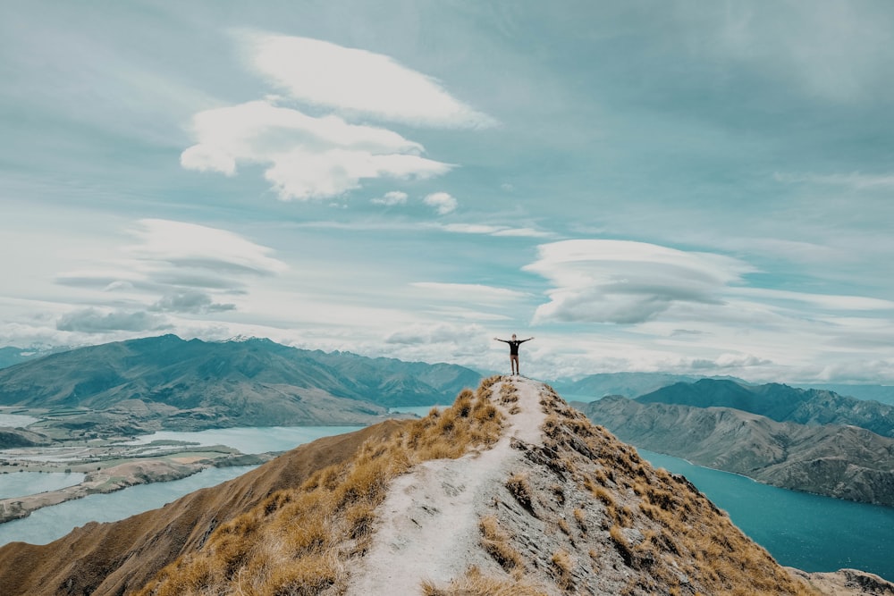 person standing on peak of the mountain near lake under white cloud blue skies