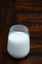 A glass of milk is placed on a wooden surface. The background consists of a dark wooden texture with defined lines and patterns. The milk in the glass appears to be fresh, with small bubbles visible on the top layer.