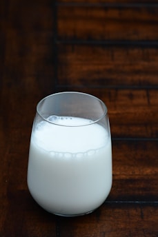 A glass of milk is placed on a wooden surface. The background consists of a dark wooden texture with defined lines and patterns. The milk in the glass appears to be fresh, with small bubbles visible on the top layer.