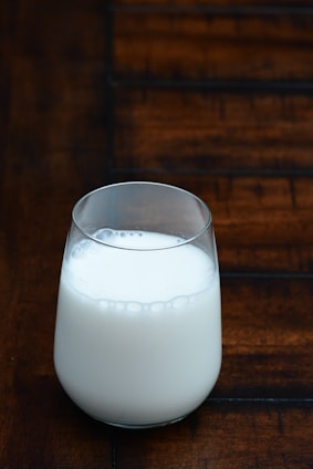 A fresh glass of creamy milk poured from a rustic jug on a wooden table with green pastures in the background.