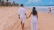 Couple walking hand-in-hand along a sandy beach with gentle waves under a clear blue sky.