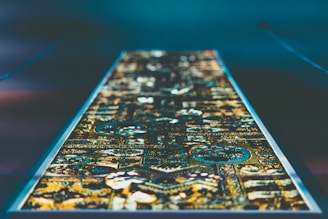 A rectangular, intricate textile pattern is laid out under dim lighting, showcasing an array of geometric shapes and detailed designs. The colors are vibrant and appear to be illuminated, giving a modern, almost digital feel to the traditional pattern.