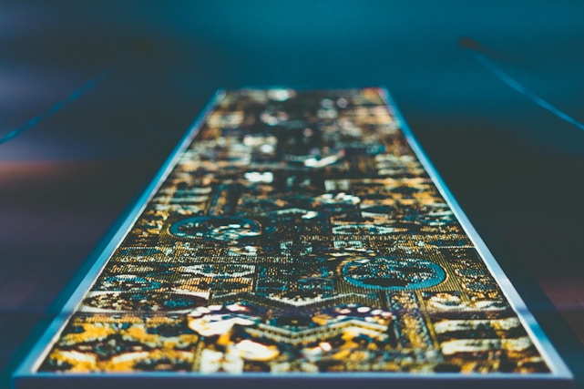 A rectangular, intricate textile pattern is laid out under dim lighting, showcasing an array of geometric shapes and detailed designs. The colors are vibrant and appear to be illuminated, giving a modern, almost digital feel to the traditional pattern.