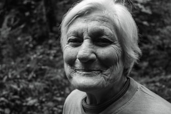 An elderly person with short white hair is smiling gently, showing facial wrinkles that suggest experience and kindness. The background features a blurred, natural setting with foliage.