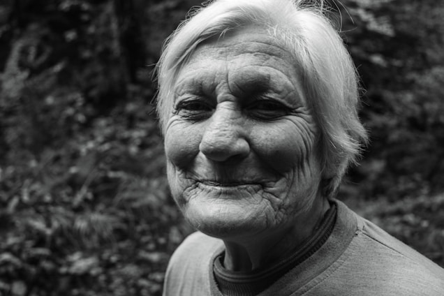 An elderly person with short white hair is smiling gently, showing facial wrinkles that suggest experience and kindness. The background features a blurred, natural setting with foliage.