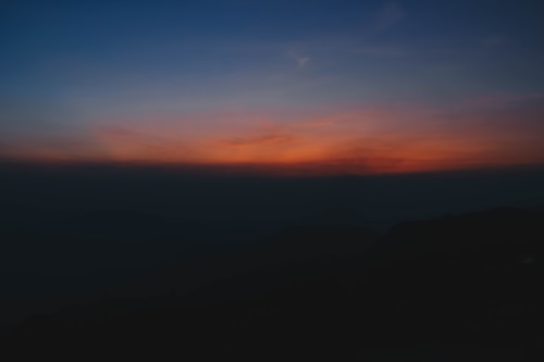 A serene landscape with a gradient sky transitioning from deep blue to vibrant orange and pink hues, giving a sense of dusk or dawn. The horizon is silhouetted against the colorful sky, with darkened landforms barely visible beneath.