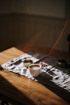 A white cup filled with coffee sits on a wooden table. The table is partially covered with a white cloth, and small decorative items are scattered around. Soft lighting creates gentle shadows, adding a warm and inviting atmosphere.